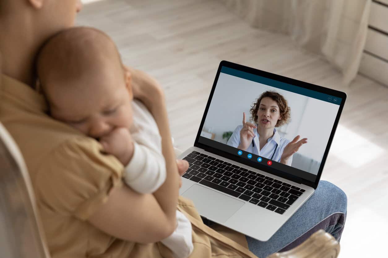 New mom holding cute napping baby in arms, making video call to doctor, consulting pediatrician from home.