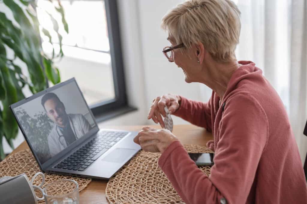 Senior woman wearing a coral sweatshirt sits at a wooden table during a telehealth appointment, gesturing while speaking to a male healthcare provider visible on her laptop screen