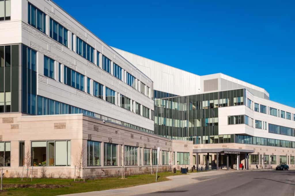 Modern multi-story healthcare facility with white exterior, floor-to-ceiling windows, and contemporary architectural design featuring curved and angular sections under a clear blue sky