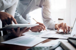 Two colleagues in button-down shirts collaborating over printed financial graphs and bar charts, with one using a laptop and another holding a tablet.